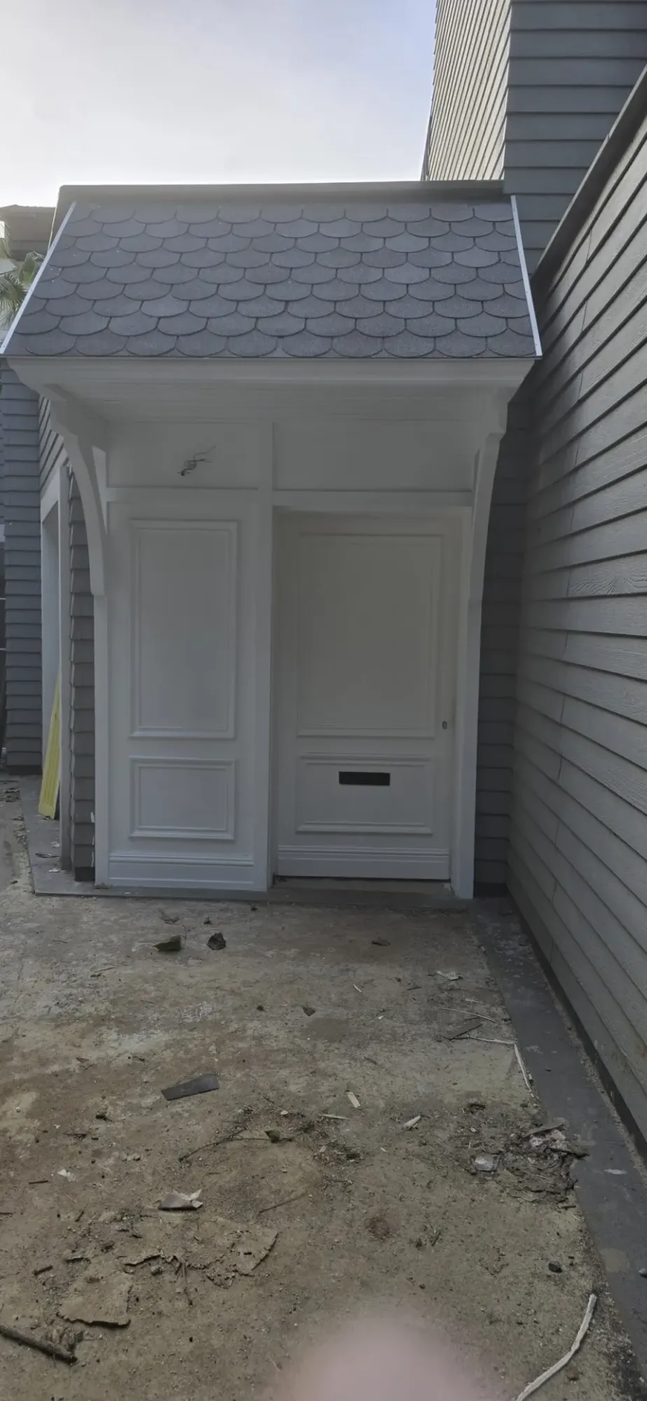 Shingled canopy and white panelled entry door between grey clapboard walls