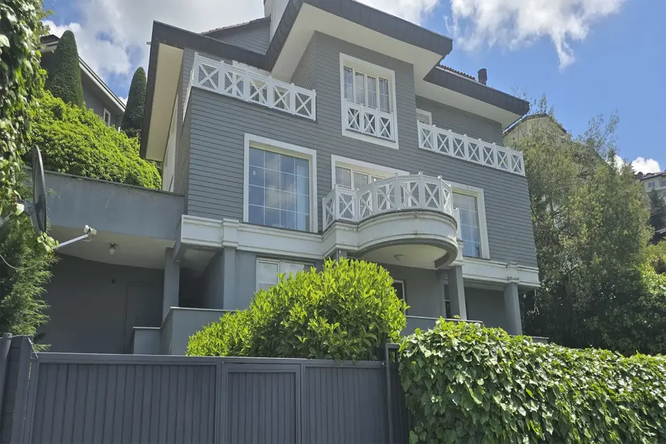 Grey villa front with a semicircular balcony behind an ivy-covered garden wall