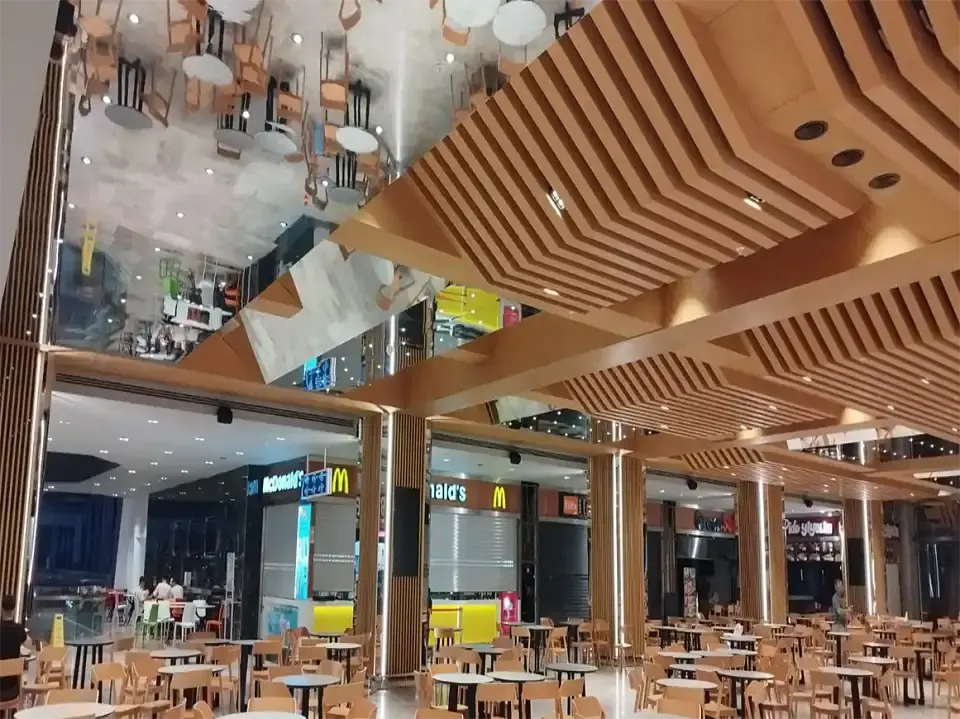 Food court with stepped wooden slatted ceiling, a mirrored ceiling panel and rows of tables and chairs